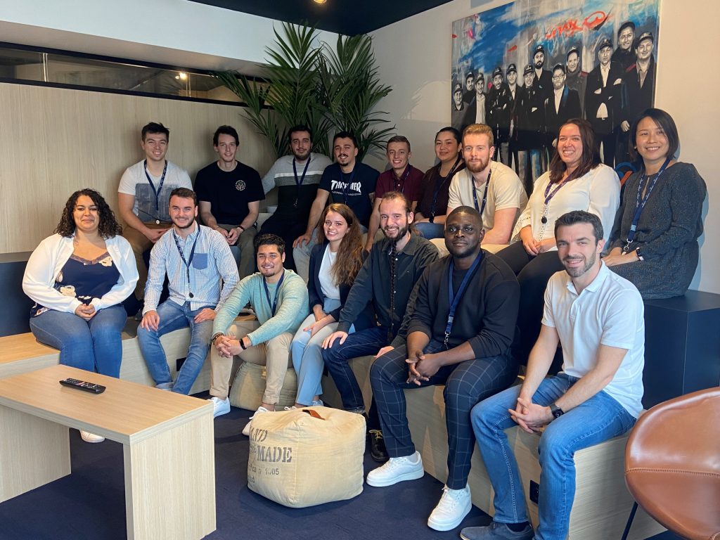 A group photo of AmeXio team members in a theater room, seated in two rows, one behind the other. There are 16 people in total.