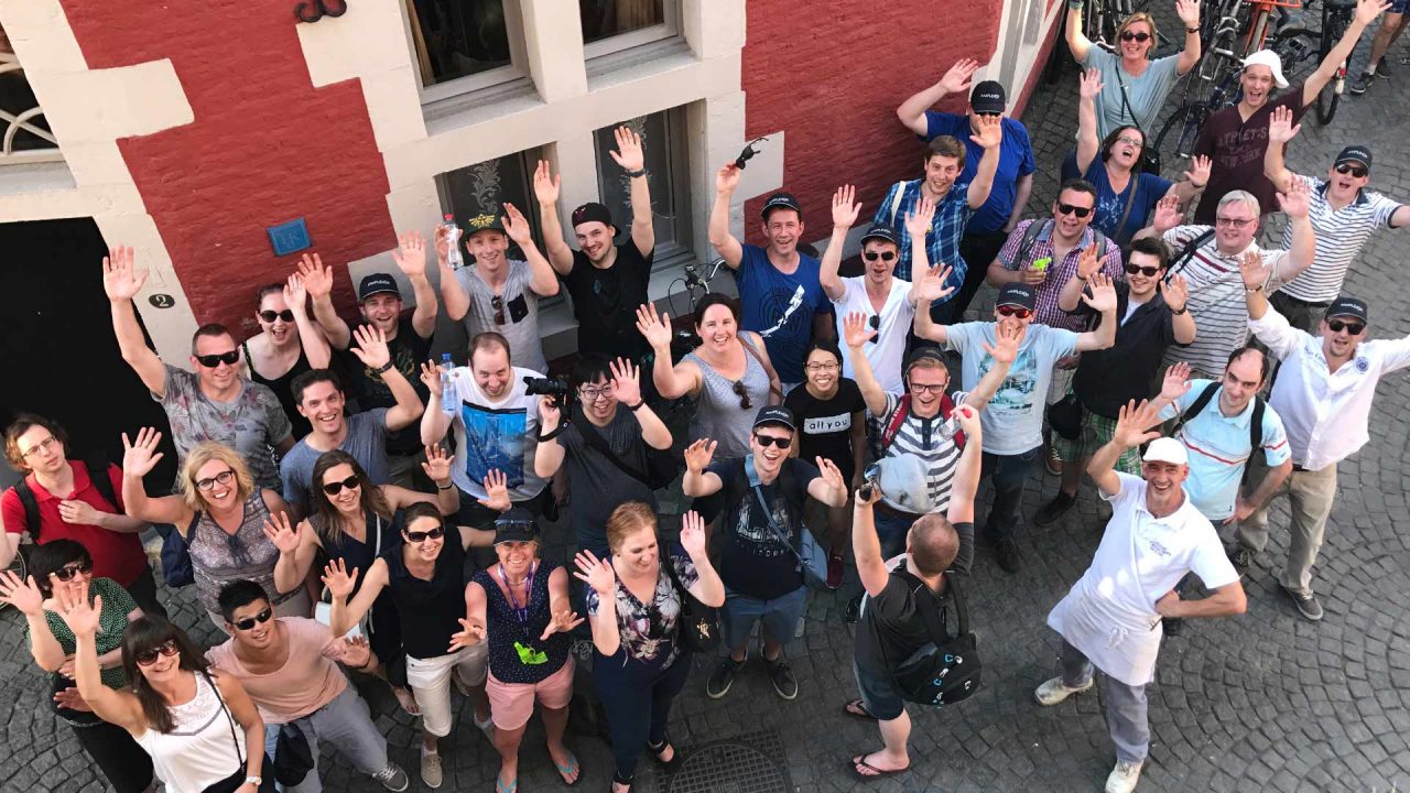 A group picture taking from above of the AmeXio Digital Support team, along with other AmeXio employees. They are standing outside on a cobbled street. There are about thirty people in the picture, and they're all smiling and throwing their hands in the air.