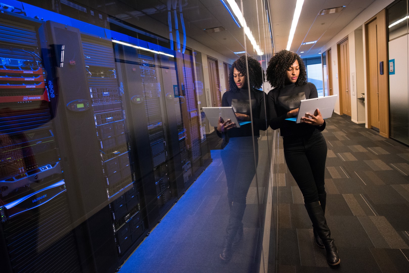 A woman leaning against the glass wall of a server room, looking at the laptop she's holding.