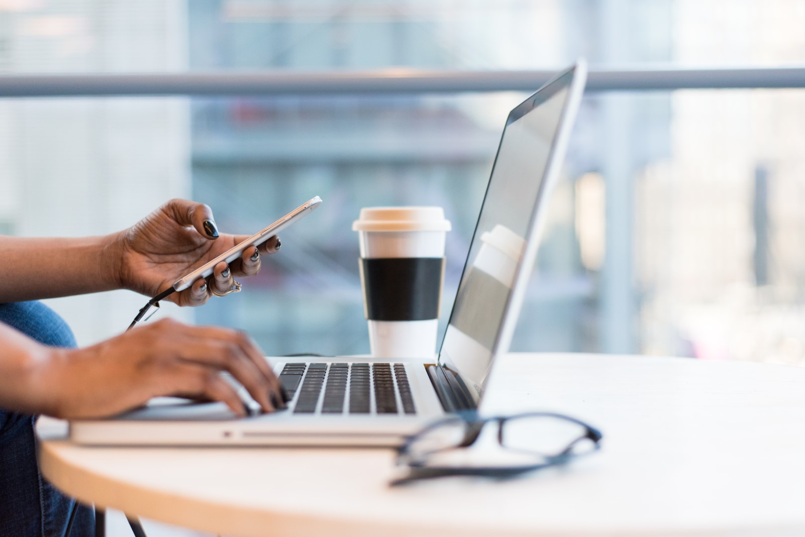 A side profile of woman holding a phone while sitting at a laptop. Only her hands are visible.