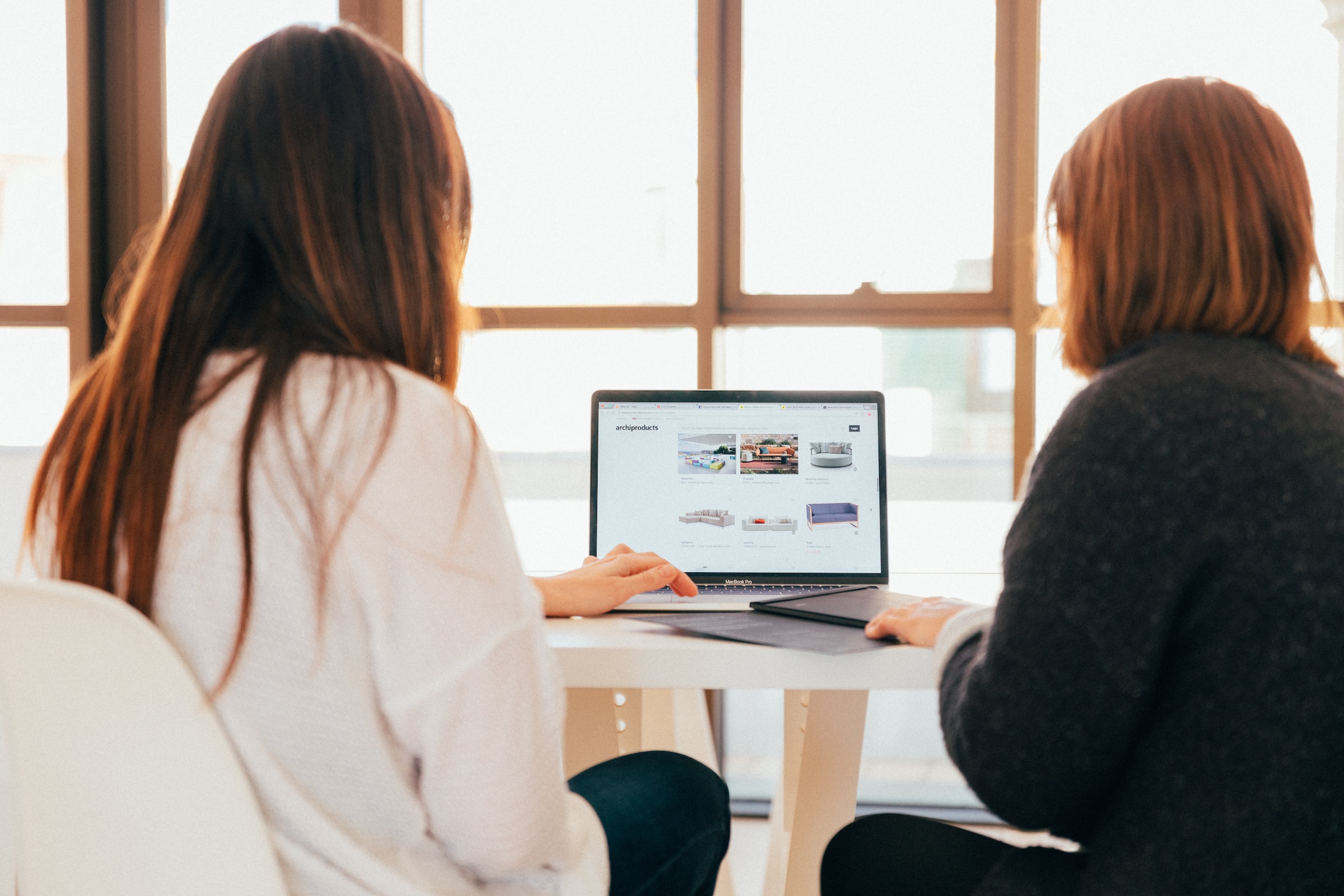 Two women seen form the back, sitting at a table and looking at the same laptop.