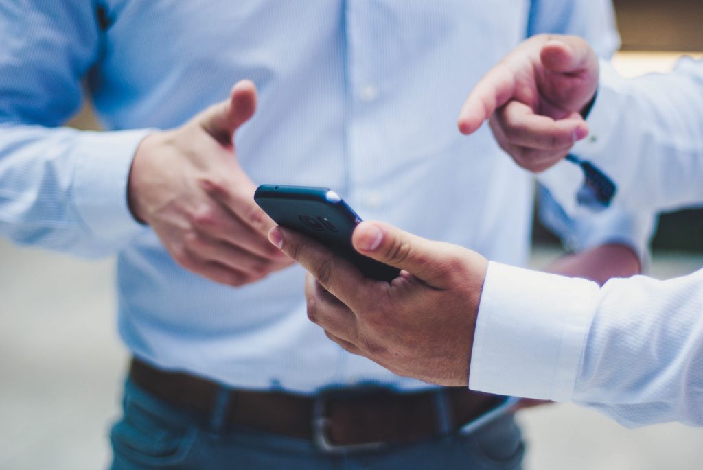 An image of a someone holding a phone, pointing at the screen and showing it to another person. They are both wearing light-colored dress shirts.