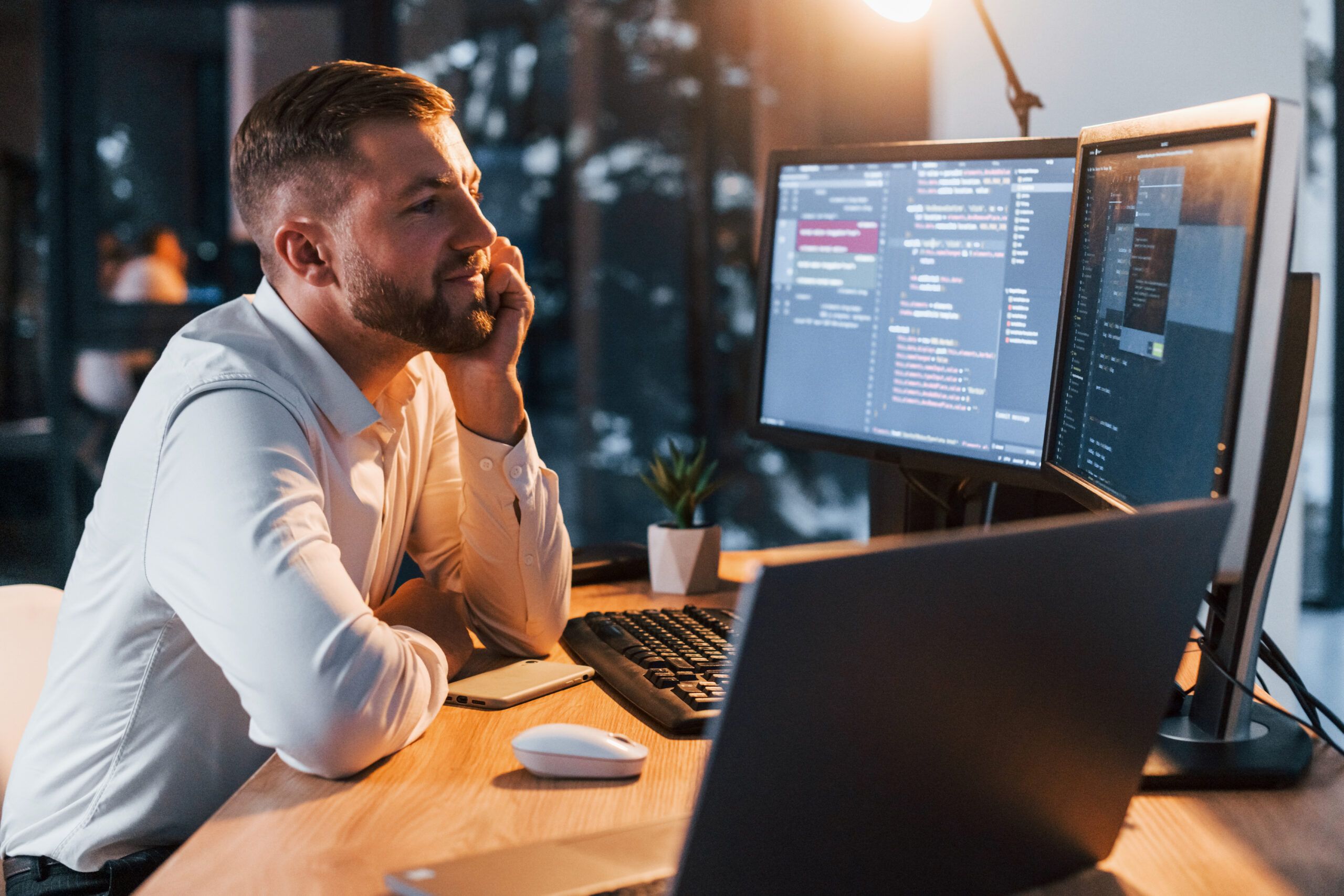 Night time. Young businessman in formal clothes is in office with multiple screens.